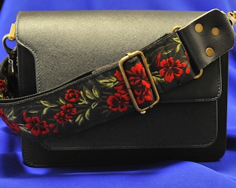 May include: A black leather handbag with a floral patterned strap. The strap is black with red flowers and green leaves. The bag has a flap closure and a gold buckle.
