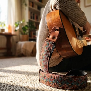 Retro woven guitar strap attached to an acoustic guitar resting in sunlight. Detailed red and teal floral embroidery adds vintage charm to a home studio decor.