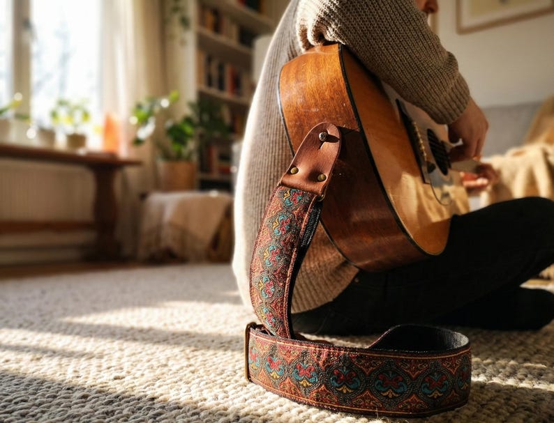Retro woven guitar strap attached to an acoustic guitar resting in sunlight. Detailed red and teal floral embroidery adds vintage charm to a home studio decor.