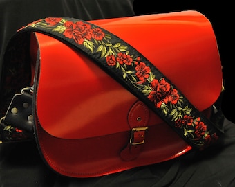 May include: A red leather satchel bag with a black and gold floral strap. The bag has a flap closure with a brass buckle.