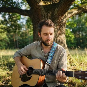 Bearded musician playing acoustic guitar under a tree with a turquoise embroidered strap. Natural vibe accessory with leather ends for comfort.