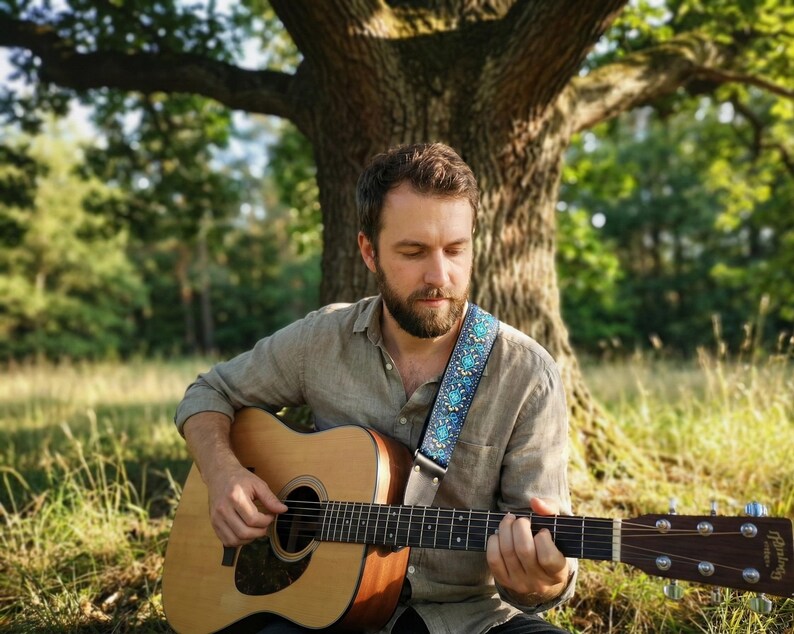 Bearded musician playing acoustic guitar under a tree with a turquoise embroidered strap. Natural vibe accessory with leather ends for comfort.
