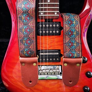 Close-up of a red electric guitar featuring an intricate red and gold jacquard strap. Durable brown leather ends secure this premium woven music accessory.