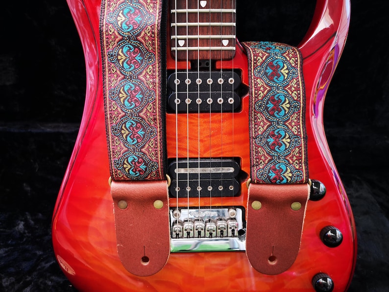 Close-up of a red electric guitar featuring an intricate red and gold jacquard strap. Durable brown leather ends secure this premium woven music accessory.
