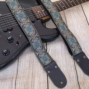 May include: A black electric guitar with a blue and gold patterned guitar strap. The strap has a leather end with a brass buckle.