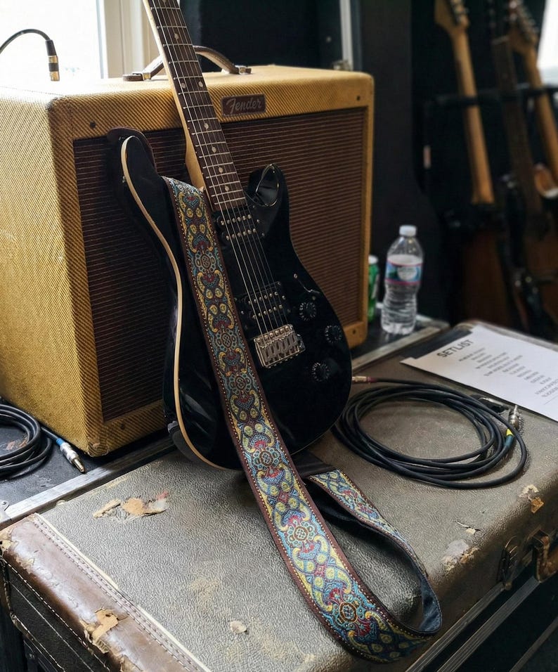Black electric guitar with a colorful paisley strap leaning against a vintage tweed amp. Stylish handmade gear setup ready for professional recording sessions.