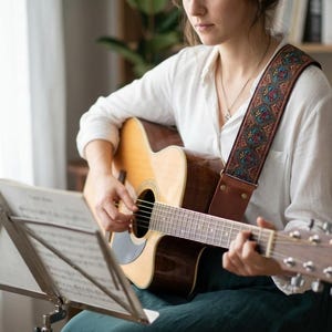 Woman playing acoustic guitar using a vintage woven strap with red and teal paisley embroidery. Comfortable boho accessory with durable leather ends for practice.