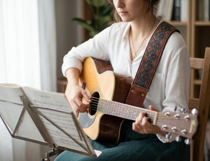 Woman playing acoustic guitar using a vintage woven strap with red and teal paisley embroidery. Comfortable boho accessory with durable leather ends for practice.