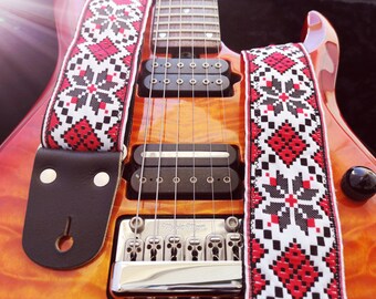 May include: A red, black, and white patterned guitar strap with a black leather end. The strap is attached to a red electric guitar with a chrome bridge and black pickups.
