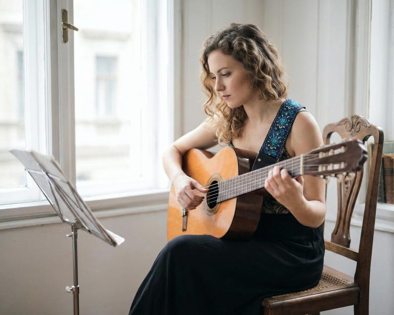 Woman playing classical guitar near a window featuring a blue floral guitar strap. Comfortable woven accessory with a boho vibe for musicians.