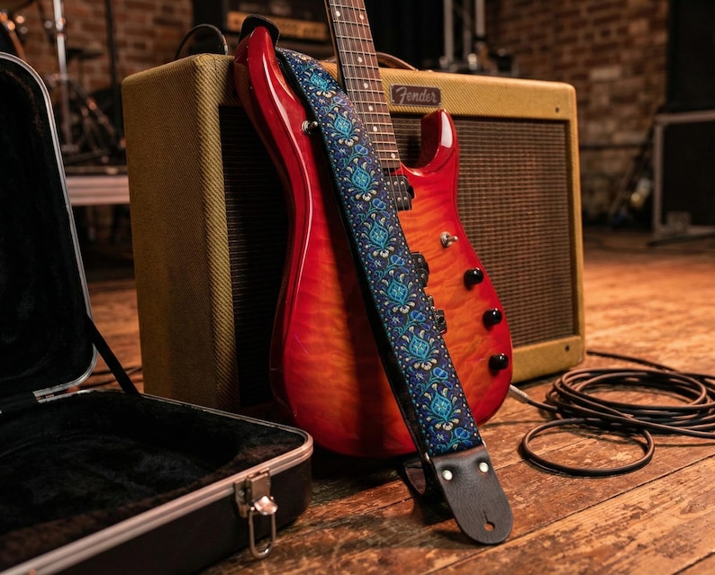Red electric guitar leaning on a tweed amp displaying a blue floral guitar strap. High-quality handmade gear with leather ends for studio setups.