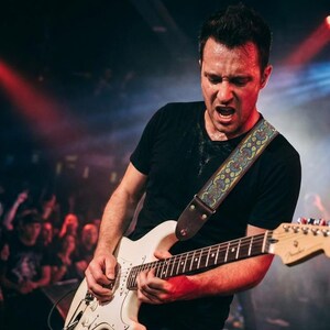 Energetic guitarist wearing a patterned woven guitar strap during a live concert with red stage lights. Durable electric guitar accessory built for intense performances.