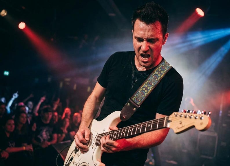 Energetic guitarist wearing a patterned woven guitar strap during a live concert with red stage lights. Durable electric guitar accessory built for intense performances.