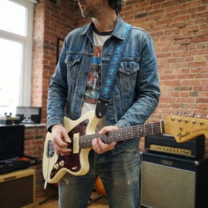 Guitarist in a denim jacket playing a white electric guitar with a blue woven strap. Vintage style accessory offering comfort for standing players.
