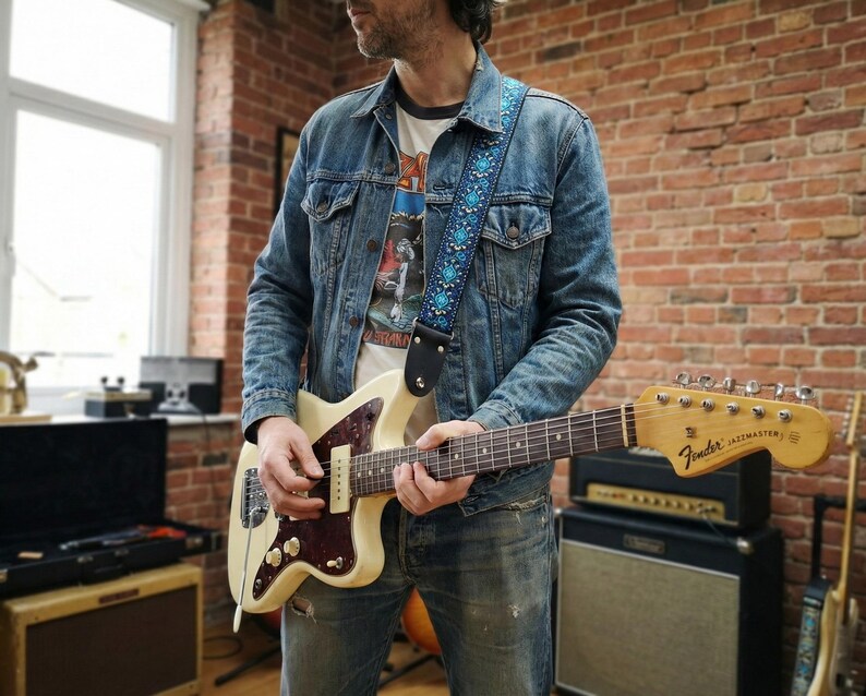 Guitarist in a denim jacket playing a white electric guitar with a blue woven strap. Vintage style accessory offering comfort for standing players.