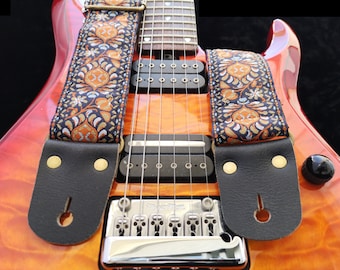 May include: A close-up of a red electric guitar with a black leather guitar strap with a colorful patterned design. The guitar has two black pickups and a chrome bridge.