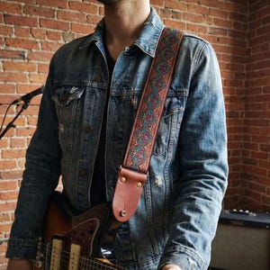 Man in denim jacket holding an electric guitar with a red and blue paisley woven strap. Stylish vintage embroidered gear perfect for gigging musicians.
