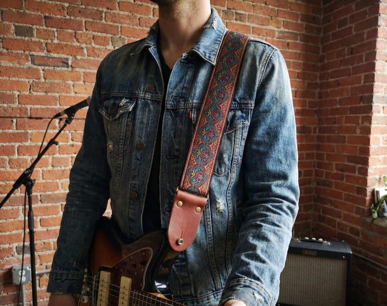 Man in denim jacket holding an electric guitar with a red and blue paisley woven strap. Stylish vintage embroidered gear perfect for gigging musicians.