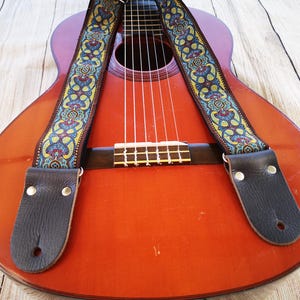 Vintage blue and gold paisley guitar strap draped over a classical acoustic guitar body. Durable woven accessory with leather ends for a stylish boho folk look.