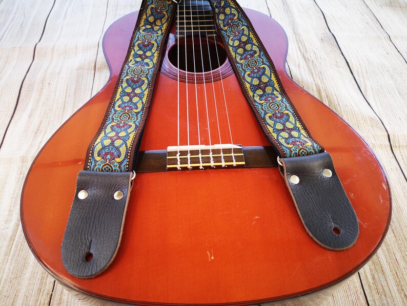 Vintage blue and gold paisley guitar strap draped over a classical acoustic guitar body. Durable woven accessory with leather ends for a stylish boho folk look.