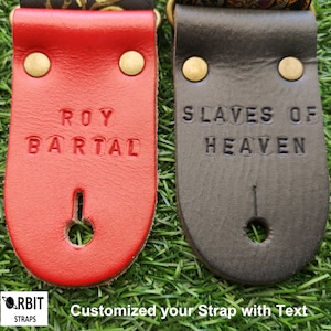 Personalized leather guitar strap ends custom stamped with names in red and black. Handmade musical accessory options displayed on green grass for gifts.