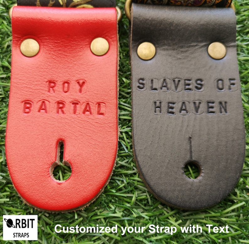Personalized leather guitar strap ends custom stamped with names in red and black. Handmade musical accessory options displayed on green grass for gifts.