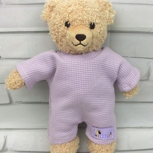 Könnte beinhalten: Ein Teddybär mit hellbraunem Fell, der einen hellvioletten Strampler trägt. Der Strampler hat eine Waffelstruktur und einen kleinen Aufnäher mit einer Dackel-Illustration. Der Bär hat eine braune Nase und schwarze Augen.