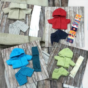 Op de afbeelding: Een collage van poppenkledingsets in verschillende kleuren. Sets bevatten jassen, shorts, broeken, sjaals en handschoenen. Kleuren zijn beige, rood, blauw, groen en wit. De kleding is gerangschikt op een houten achtergrond.