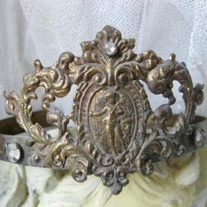 Pode incluir: Uma coroa de metal em tom dourado com um design de filigrana ornamentado e um medalhão oval central que retrata uma figura. A banda é acentuada com cristais transparentes de corte redondo incrustados no topo central e nas laterais.