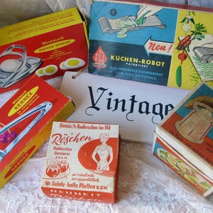 Peut inclure: Une collection de cinq boîtes d'ustensiles de cuisine vintage avec des illustrations graphiques colorées et du texte en allemand. Les boîtes incluent Westmark Duplex-Spezial, Kuchen-Robot, Kernex, Röschen et Eier-Stückler, disposées autour d'un panneau blanc avec le mot Vintage.