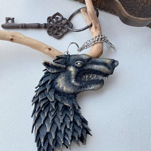May include: A detailed pendant featuring a wolf's head with intricate, layered detailing in shades of black and gold. The pendant hangs from a silver chain. An antique key and a vintage leather case are in the background.
