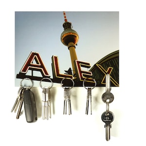 May include: A key holder with a photo of the Berlin TV Tower and the word "ALEX" in neon lights. Five sets of keys are hanging from the holder.