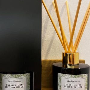 May include: A black glass reed diffuser with a gold collar and wooden reeds. The label reads "Smith & Greening FRESH LINEN REED DIFFUSER Vegan & Cruelty Free HANDMADE IN YORKSHIRE 100ML". A black box is on the left.