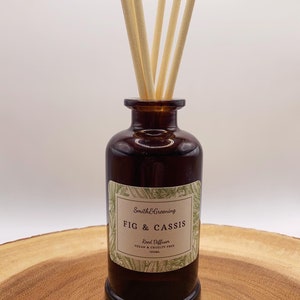 May include: A brown glass bottle with a label that reads "Smith & Greening FIG & CASSIS Reed Diffuser VEGAN & CRUELTY FREE 100ML". The bottle has wooden sticks inserted into the top.