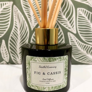 May include: A black glass bottle with a gold lid and a label that reads "Smith & Greening FIG & CASSIS Reed Diffuser VEGAN & CRUELTY FREE 100ML". The bottle is filled with a clear liquid and has wooden sticks sticking out of the top.
