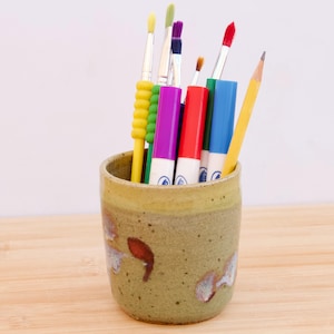May include: A speckled, olive-green ceramic cup contains art supplies. It holds paintbrushes with yellow, purple, and red bristles, a yellow pencil, and colourful markers. The cup is on a wooden surface, with a light background.