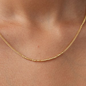 May include: A close-up of a person wearing a gold chain necklace. The chain is thin and delicate, and it is made of small, round beads.