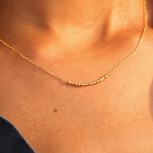 May include: A fine gold necklace featuring a series of tiny beads on a delicate chain. The necklace is displayed against a dark blue shirt. The image is a close-up, showcasing the jewellery's minimalist design.