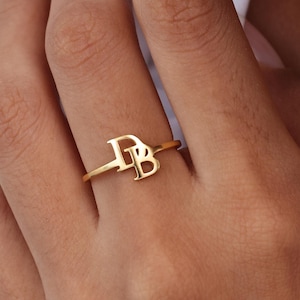 May include: A gold ring displaying the intertwined letters "D" and "B". The ring is worn on a finger, with the letters as the focal point. The band is slender, and the design is elegant and minimalist. The ring is likely made of gold.
