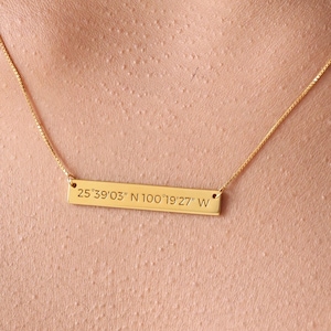 May include: A gold necklace with a thin chain and a rectangular pendant engraved with the coordinates "25.3903° N 100.1927° W".
