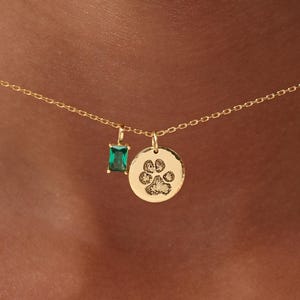 May include: A gold necklace featuring two pendants. One is a small, rectangular emerald-green gemstone. The other is a circular charm with a paw print design. The necklace is held up by a person.