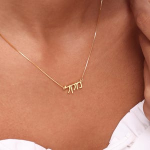 May include: A fine gold necklace with a pendant bearing Hebrew script. The necklace is set against a white blouse. The pendant is the central element, highlighting the detailed design and craftsmanship. The necklace is a simple yet elegant accessory.