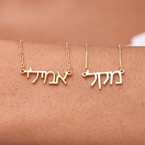 May include: Two gold-colored nameplate necklaces with delicate chains. The necklaces feature Hebrew script. The necklaces are displayed on a person's arm, with a white shirt in the background.