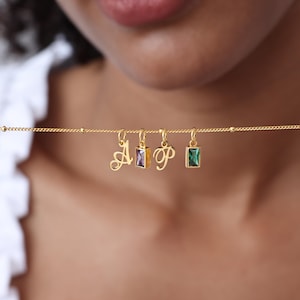 May include: A gold necklace with four charms: a cursive letter "A", a rectangular purple gem, a cursive letter "P", and a rectangular green gem. The necklace is worn around the neck, with the charms hanging below.
