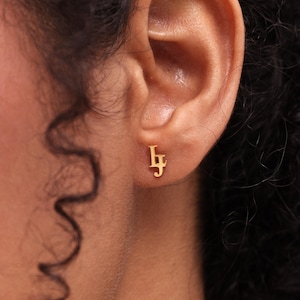 May include: A close-up of an ear with a gold-coloured earring. The earring is a small stud with the letters "lf" in a stylized font. The ear is in focus, with the earring clearly visible.