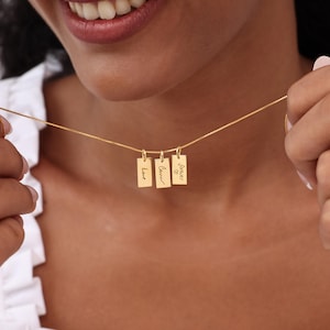 May include: A gold necklace featuring three rectangular pendants. Each pendant is inscribed with the words "Love", "Care", and "Forever" with a heart symbol. The necklace is held up by a person.
