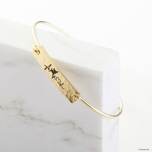 May include: Gold bangle bracelet with a hammered metal bar charm engraved with a heartbeat and the word "My Soul".