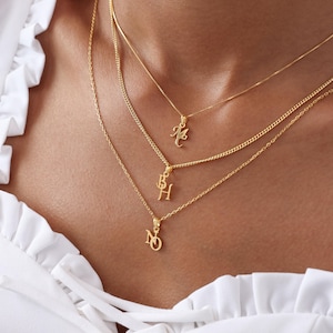 May include: A close-up of three gold necklaces with initial pendants. The necklaces are layered, with each chain at a different length. The pendants feature the letters M, BH, and NQ. The background is a person's chest and a white blouse.