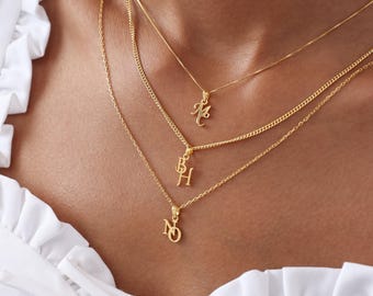 May include: A close-up of three gold necklaces with initial pendants. The necklaces are layered, with each chain at a different length. The pendants feature the letters M, BH, and NQ. The background is a person's chest and a white blouse.