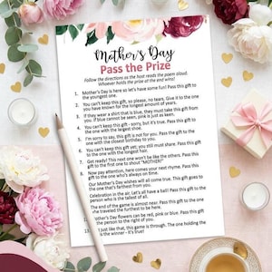 May include: A printed Mother's Day game sheet titled "Mother's Day Pass the Prize" with instructions and prompts. The sheet is surrounded by floral decorations and a pencil. A gift box and a cup of tea are also visible.